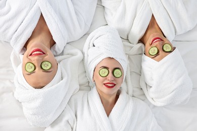 Happy friends in bathrobes with cucumber slices on bed, top view. Spa party