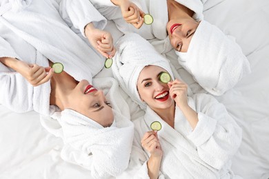 Happy friends in bathrobes with cucumber slices on bed, top view. Spa party