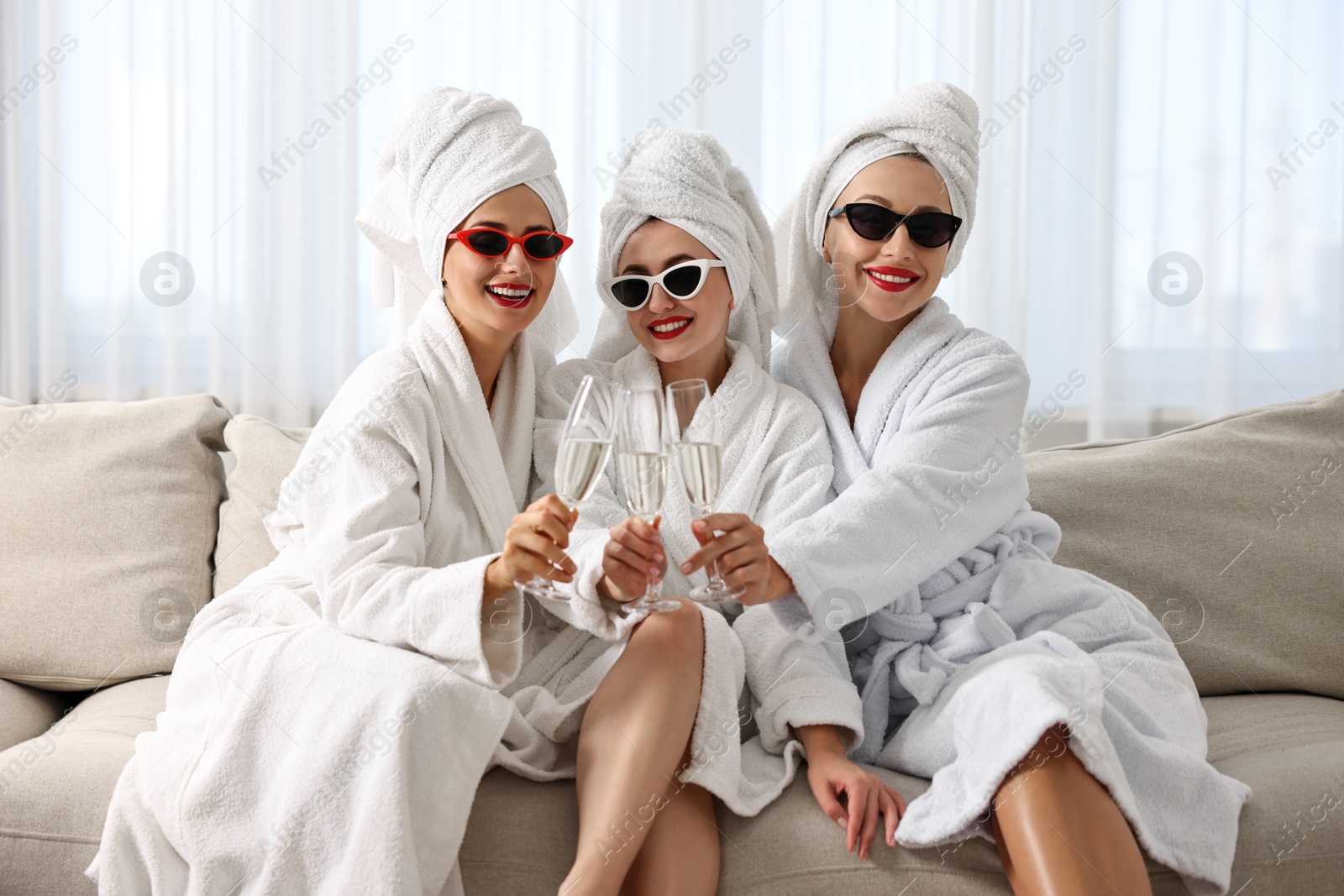 Photo of Happy friends in bathrobes with sunglasses and glasses of sparkling wine indoors. Spa party