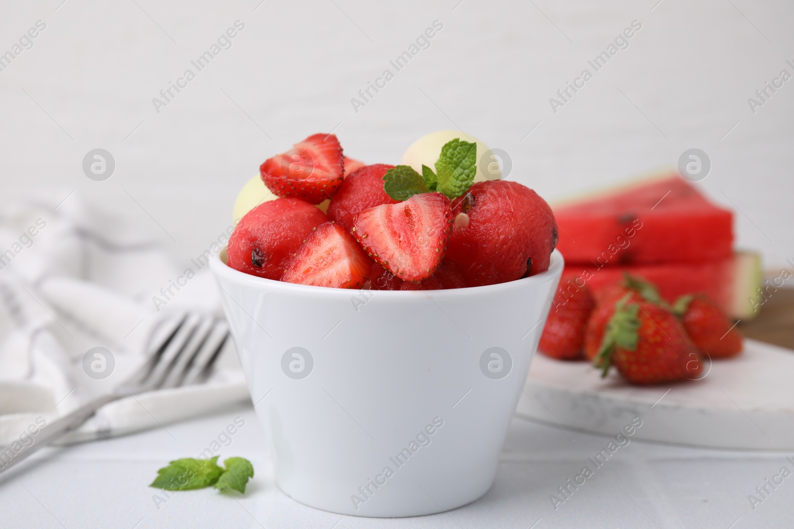 Photo of Tasty watermelon and melon balls with strawberries in bowl on white tiled table, closeup