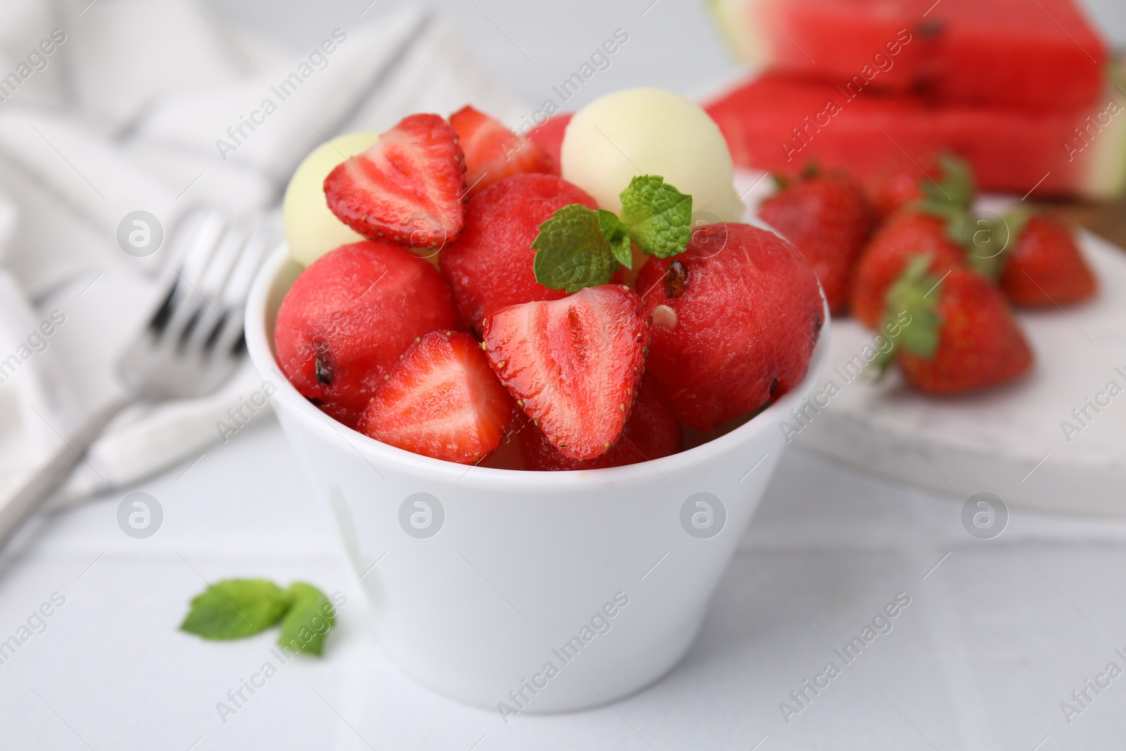 Photo of Tasty watermelon and melon balls with strawberries in bowl on white tiled table, closeup