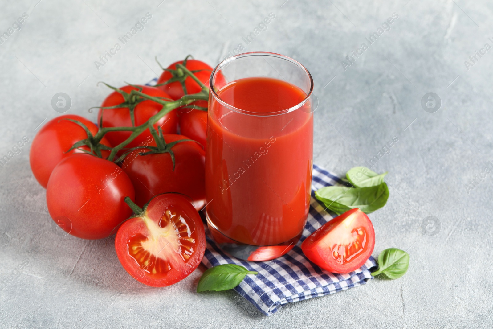 Photo of Tasty tomato juice in glass, basil leaves and fresh vegetables on grey textured table