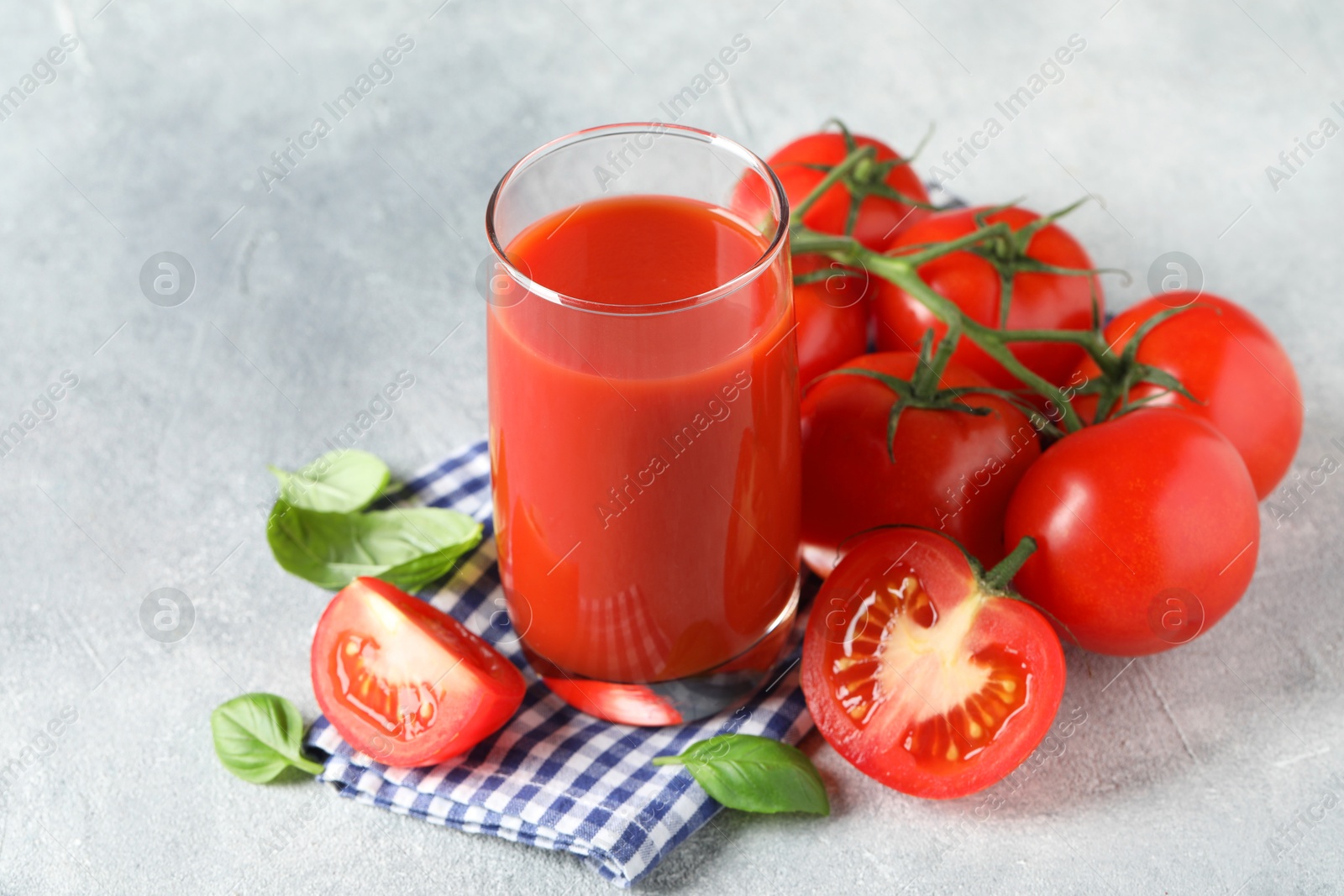 Photo of Tasty tomato juice in glass, basil leaves and fresh vegetables on grey textured table