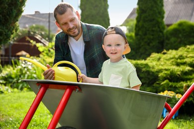 Photo of Father with watering can and his son sitting in wheelbarrow outdoors, selective focus