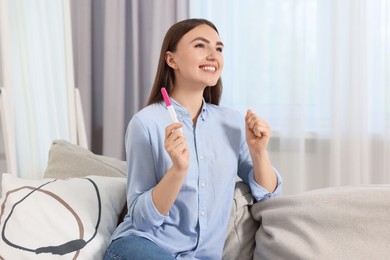 Photo of Happy woman holding pregnancy test on sofa indoors