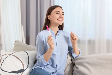 Happy woman holding pregnancy test on sofa indoors