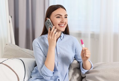 Photo of Happy woman with pregnancy test talking on smartphone indoors