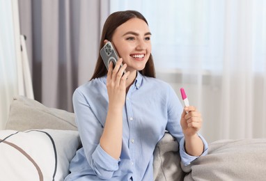 Happy woman with pregnancy test talking on smartphone indoors