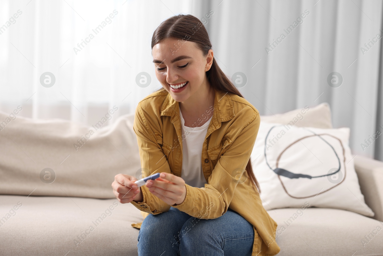 Photo of Happy woman holding pregnancy test on sofa indoors