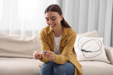 Photo of Happy woman holding pregnancy test on sofa indoors