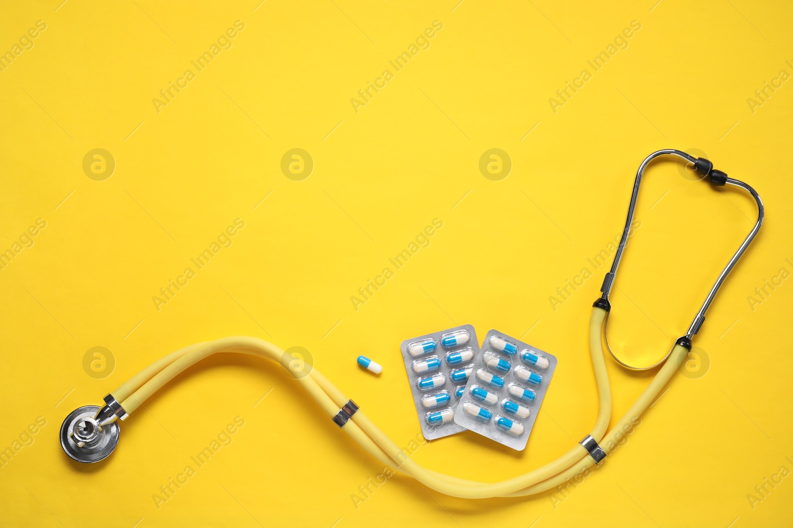 Photo of Antibiotic pills in blisters and stethoscope on yellow background, top view. Space for text