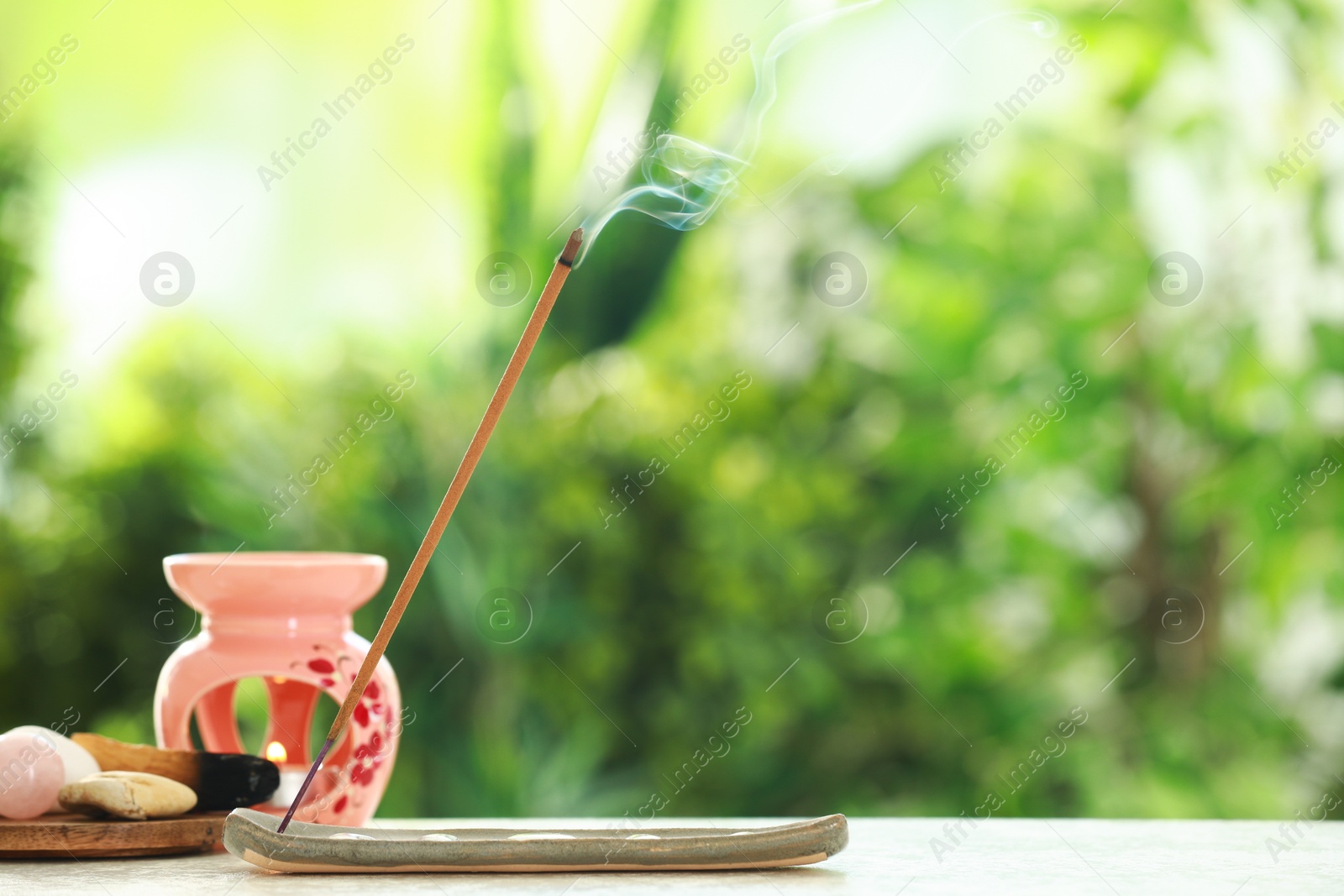 Photo of Incense stick smoldering in holder and stones on light table outdoors, space for text