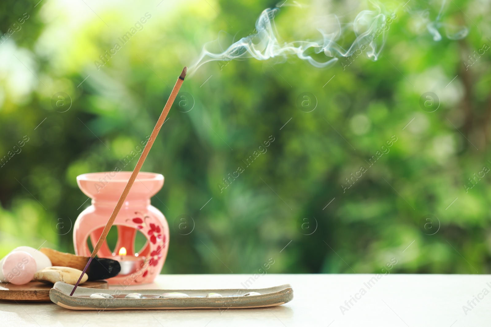 Photo of Incense stick smoldering in holder and stones on light table outdoors, space for text