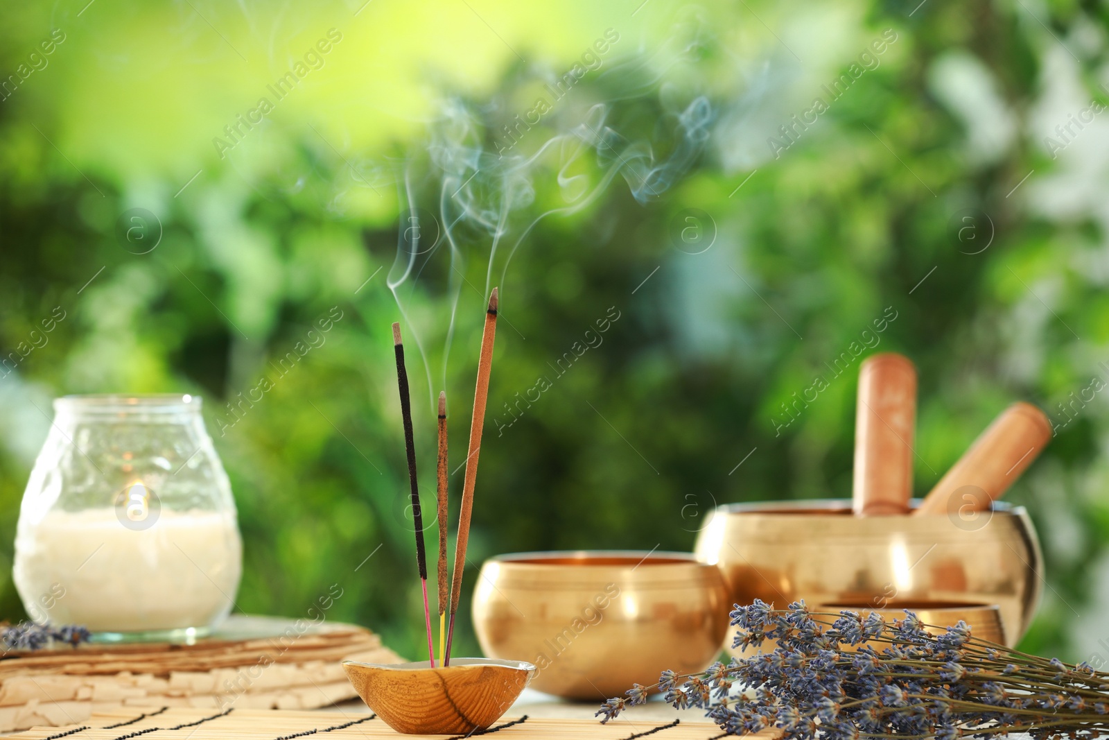 Photo of Incense sticks smoldering in holder, Tibetan singing bowls, dried lavender and candle on table outdoors