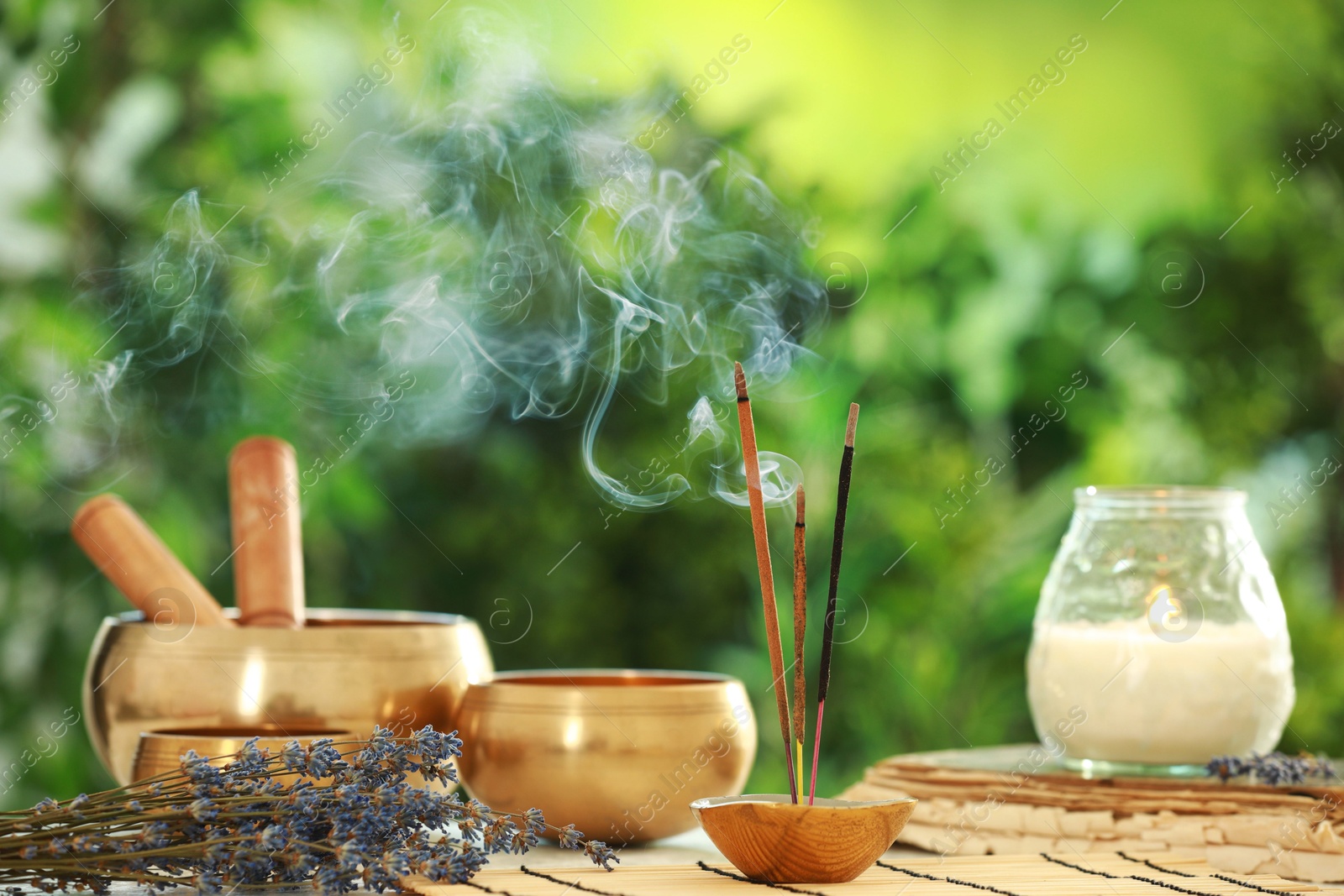 Photo of Incense sticks smoldering in holder, Tibetan singing bowls, dried lavender and candle on table outdoors