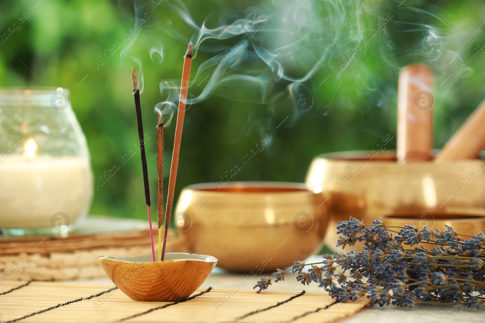 Photo of Incense sticks smoldering in holder, Tibetan singing bowls, dried lavender and candle on table outdoors