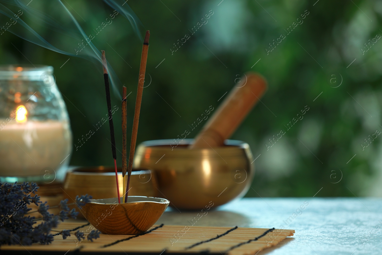 Photo of Incense sticks smoldering in holder, Tibetan singing bowls, dried lavender and candle on table outdoors