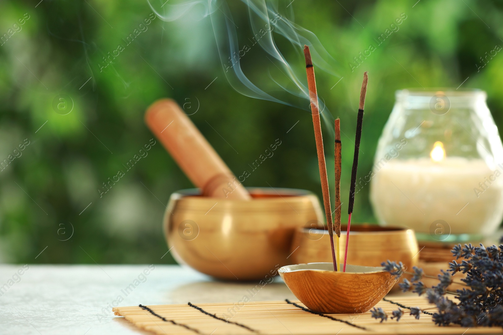 Photo of Incense sticks smoldering in holder, Tibetan singing bowls, dried lavender and candle on light table outdoors