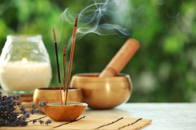 Photo of Incense sticks smoldering in holder, Tibetan singing bowls, dried lavender and candle on light table outdoors