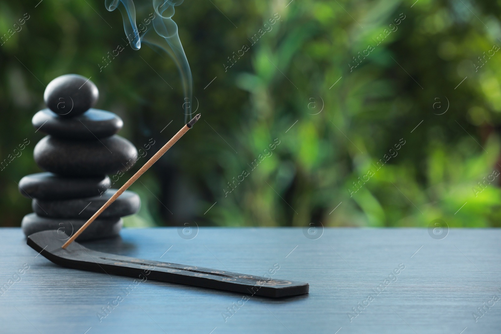 Photo of Incense stick smoldering in holder and spa stones on wooden table outdoors, space for text. Om ligature