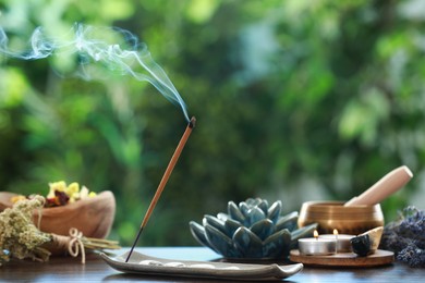 Incense stick smoldering in holder, burning candles, dry flowers and Tibetan singing bowl on wooden table outdoors