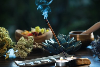 Photo of Incense stick smoldering in holder, candles and dry flowers on wooden table outdoors