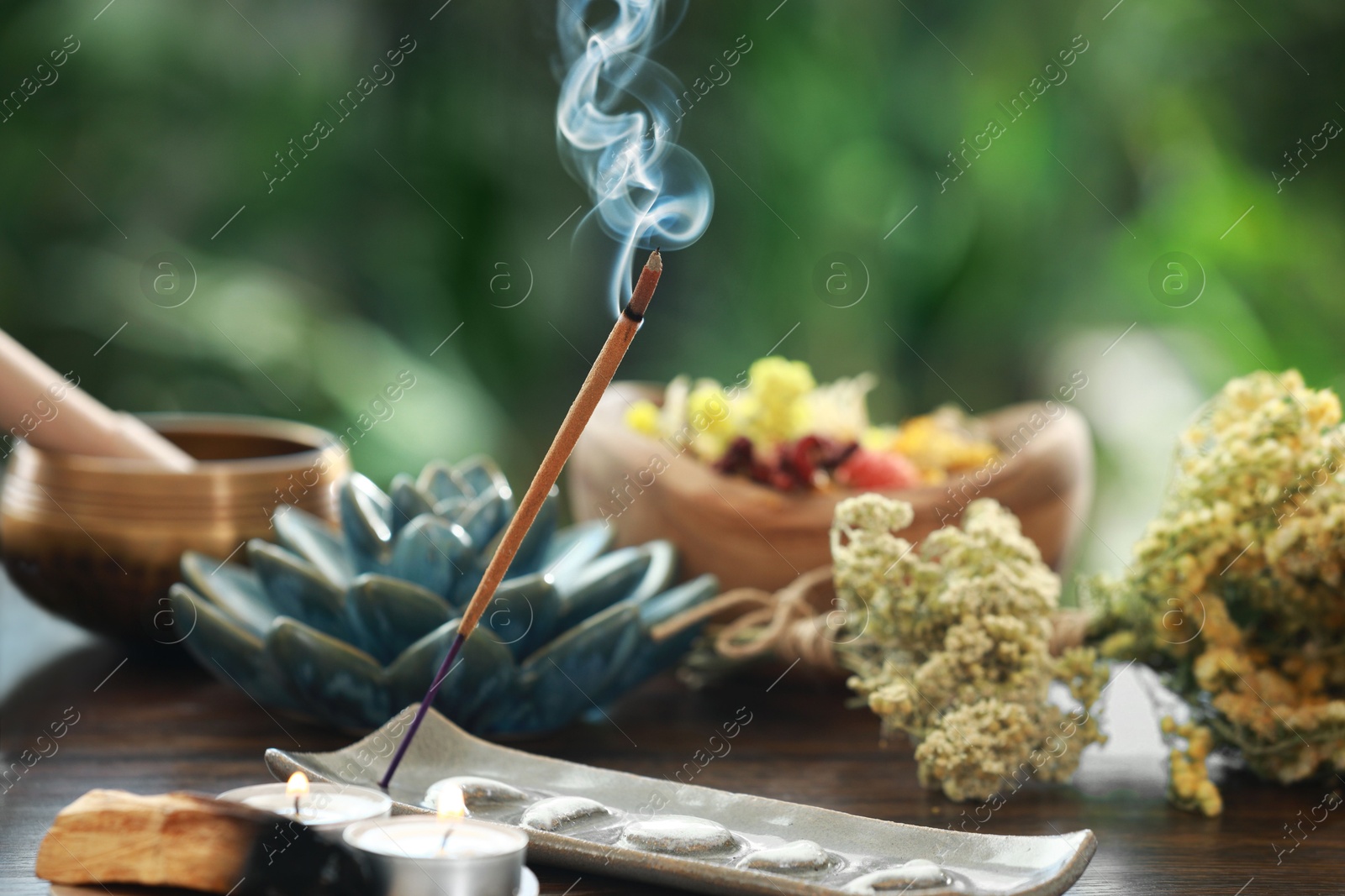 Photo of Incense stick smoldering in holder, candles and dry flowers on wooden table outdoors