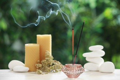 Photo of Incense sticks smoldering in holder near stones, candles and dry flowers on wooden table outdoors