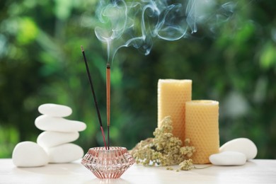 Incense sticks smoldering in holder near stones, candles and dry flowers on wooden table outdoors