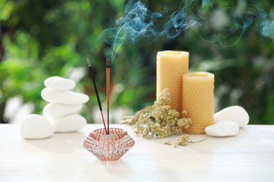 Incense sticks smoldering in holder near stones, candles and dry flowers on wooden table outdoors