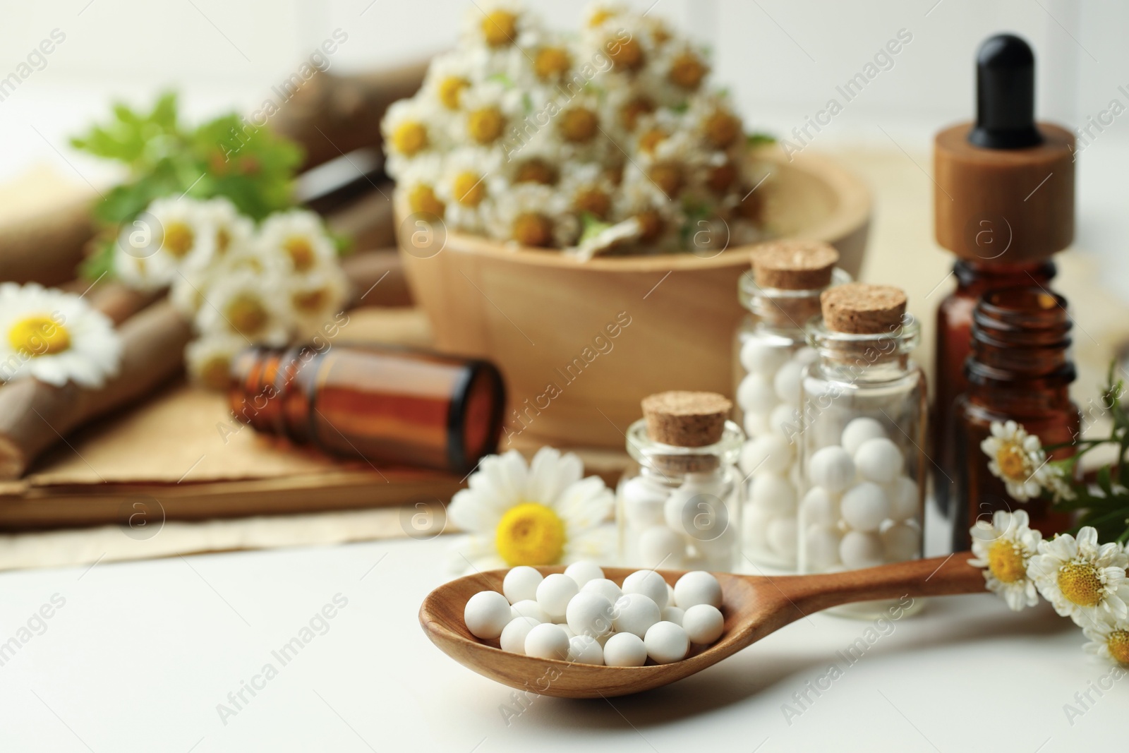 Photo of Homeopathic remedy. Spoon with pills, bottles and chamomile flowers on white table, closeup