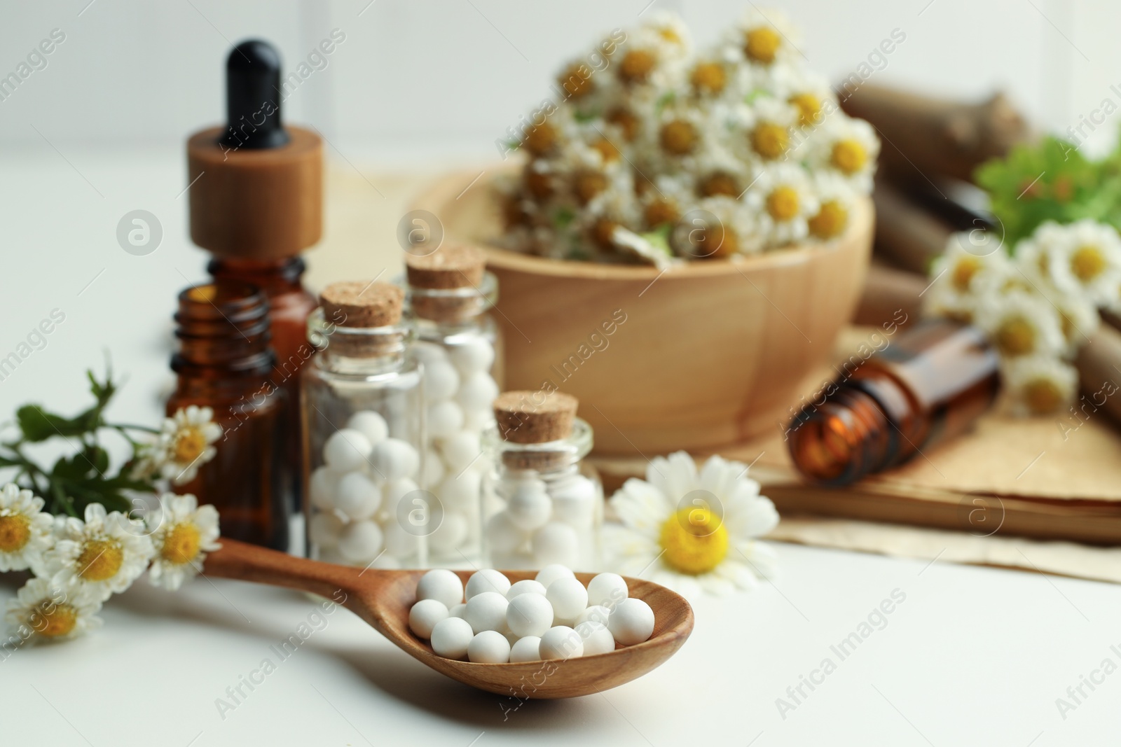 Photo of Homeopathic remedy. Spoon with pills, bottles and chamomile flowers on white table, closeup