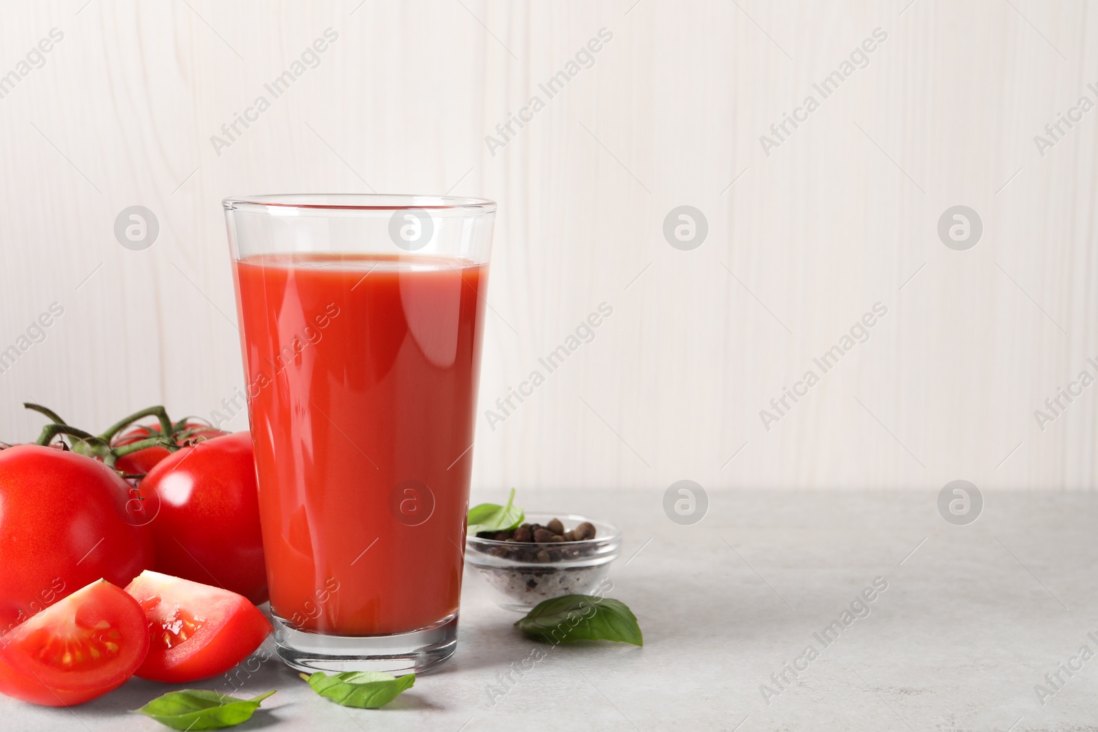 Photo of Tasty tomato juice in glass, basil leaves, peppercorns and fresh vegetables on light grey table, space for text