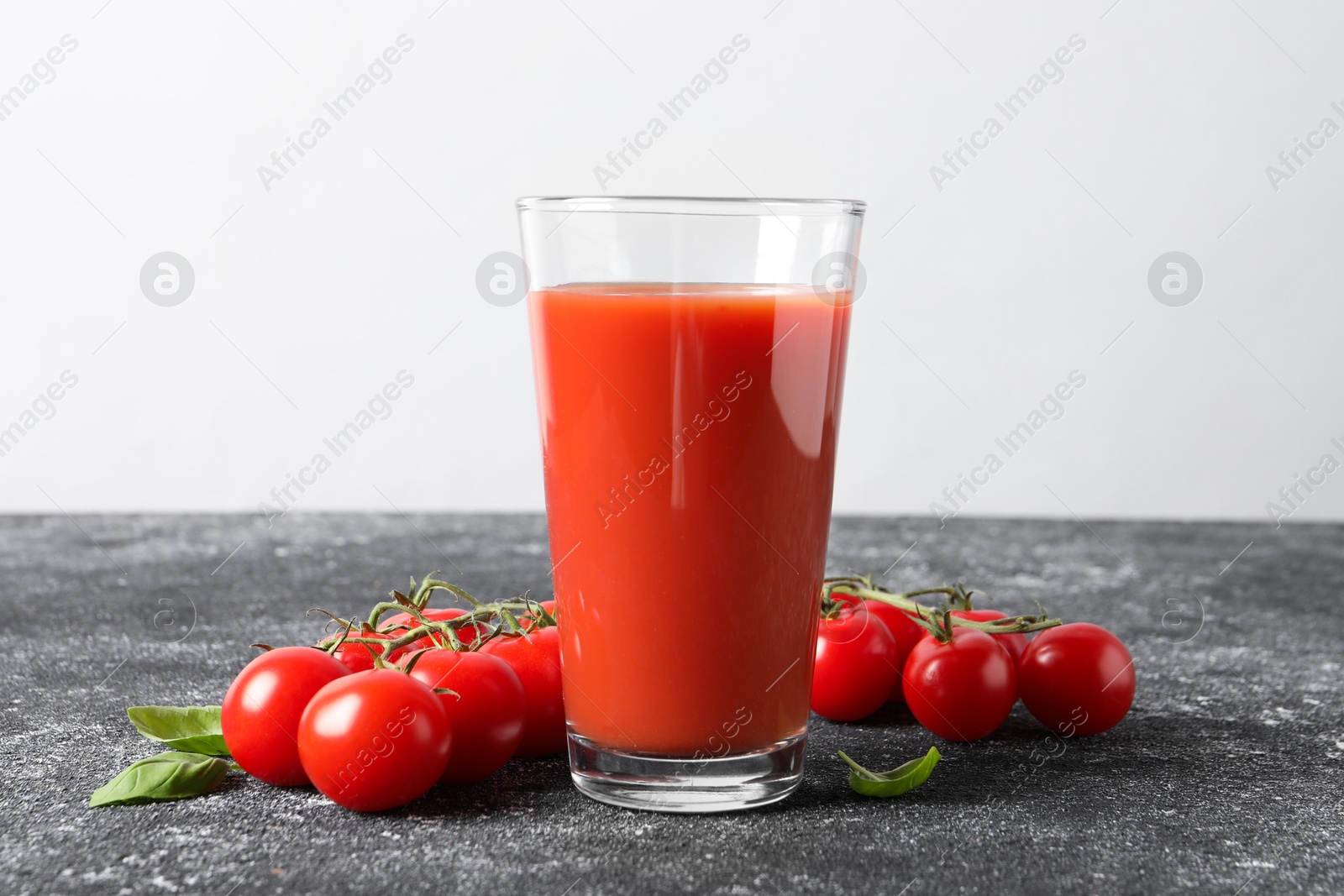 Photo of Tasty tomato juice in glass, basil and fresh vegetables on grey table