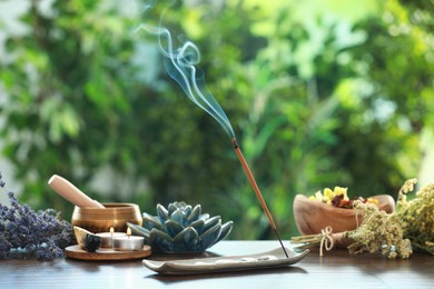 Incense stick smoldering in holder, burning candles, dry flowers and Tibetan singing bowl on wooden table outdoors