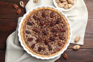 Delicious pecan pie in baking dish and fresh nuts on wooden table, flat lay