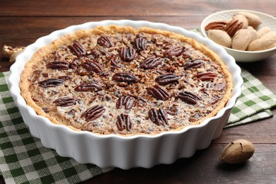 Delicious pecan pie in baking dish and fresh nuts on wooden table, closeup