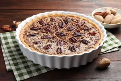 Photo of Delicious pecan pie in baking dish and fresh nuts on wooden table, closeup