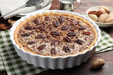 Photo of Delicious pecan pie in baking dish and fresh nuts on wooden table, closeup