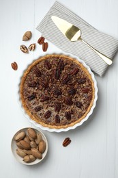 Photo of Delicious pecan pie in baking dish, fresh nuts and cake server on white wooden table, flat lay