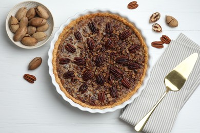 Photo of Delicious pecan pie in baking dish, fresh nuts and cake server on white wooden table, flat lay