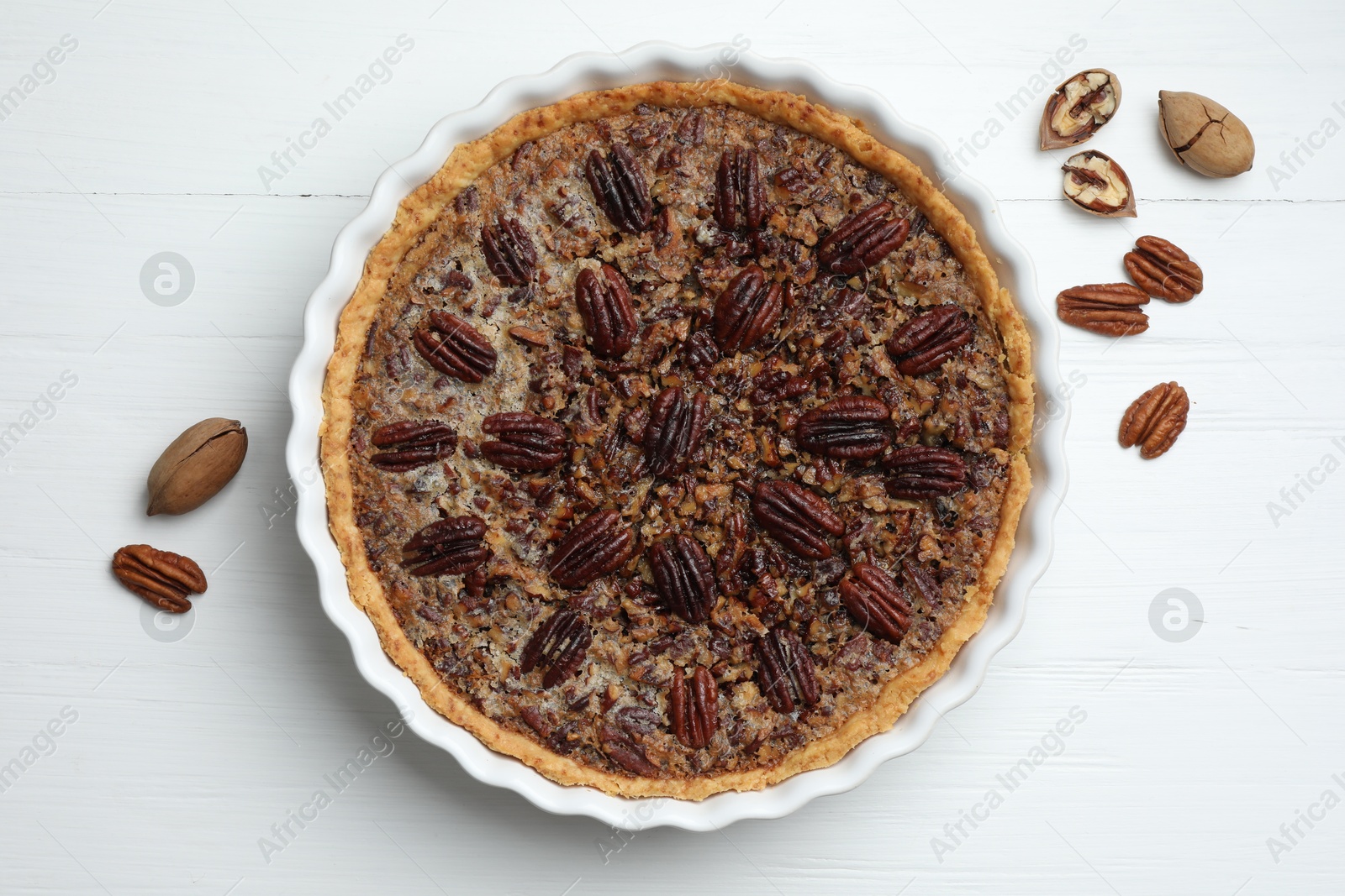 Photo of Delicious pecan pie in baking dish and fresh nuts on white wooden table, top view