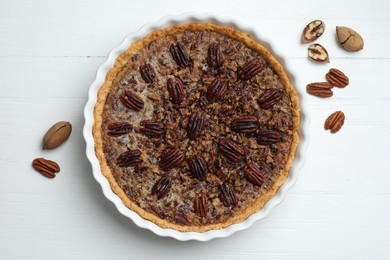 Delicious pecan pie in baking dish and fresh nuts on white wooden table, top view