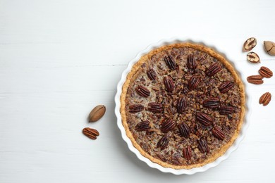 Delicious pecan pie in baking dish and fresh nuts on white wooden table, top view. Space for text