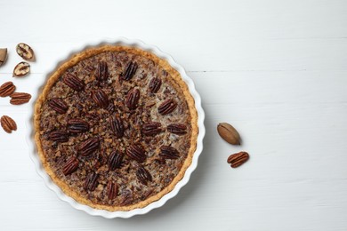 Photo of Delicious pecan pie in baking dish and fresh nuts on white wooden table, top view. Space for text