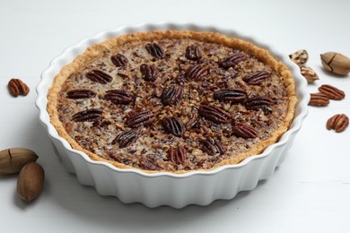 Photo of Delicious pecan pie in baking dish and fresh nuts on white wooden table, closeup