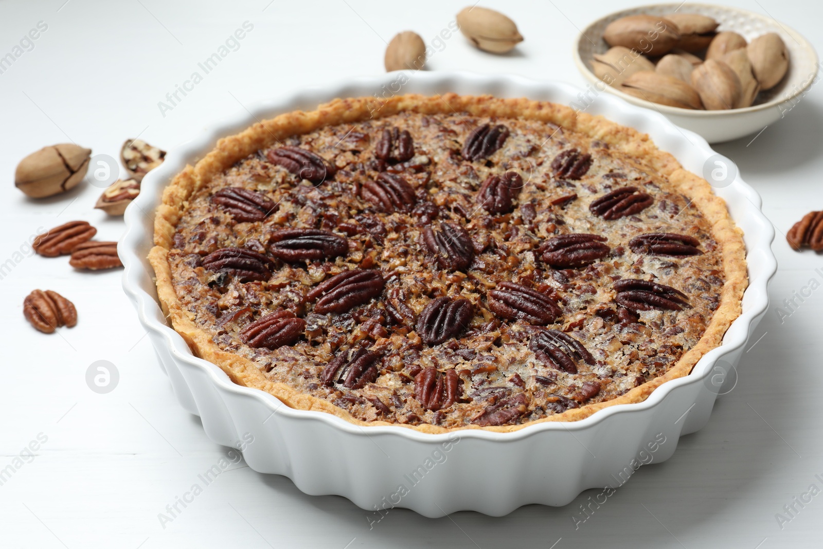 Photo of Delicious pecan pie in baking dish and fresh nuts on white wooden table, closeup