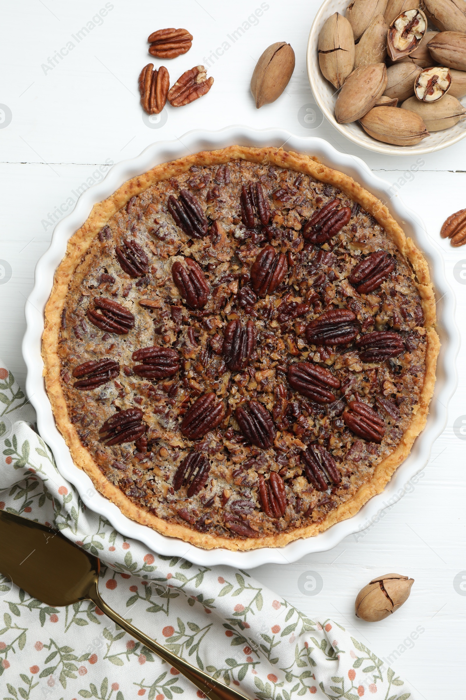 Photo of Delicious pecan pie in baking dish, cake server and fresh nuts on white wooden table, flat lay