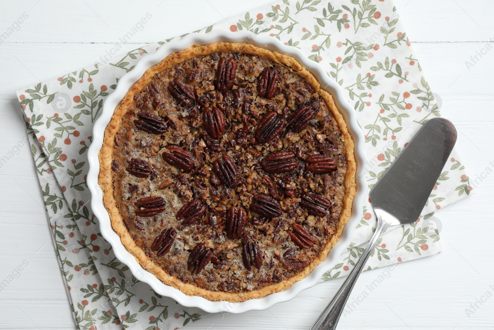 Photo of Delicious pecan pie in baking dish and cake server on white wooden table, flat lay