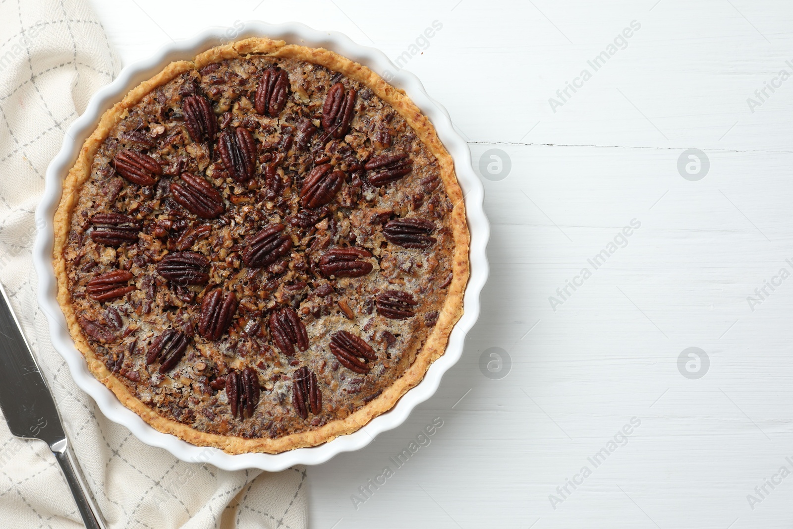 Photo of Delicious pecan pie in baking dish and knife on white wooden table, flat lay. Space for text